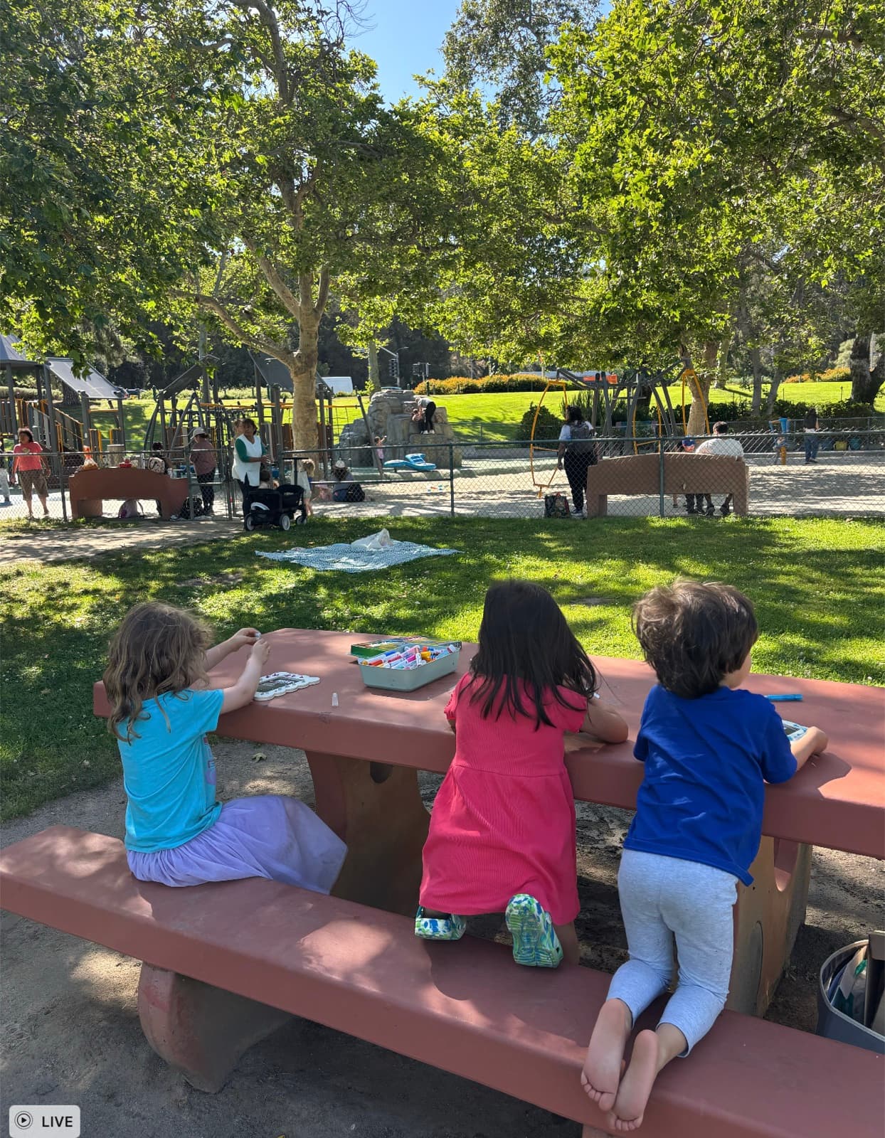 Kids at a park picnic table working on wood-frame art crafts with a playground in the background