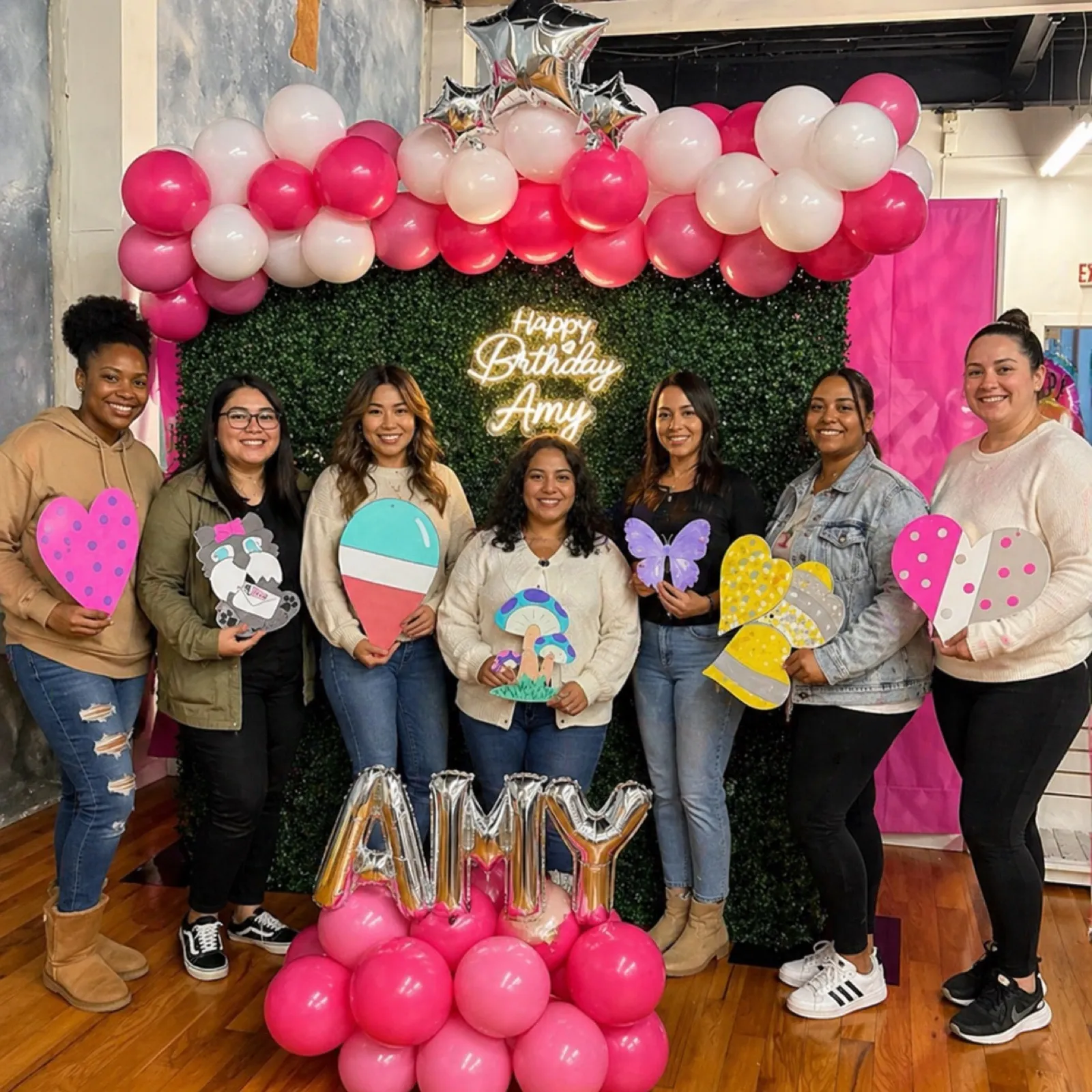 A group of coworkers celebrating with handmade paper-craft hearts and butterflies under a balloon arch at an office art party