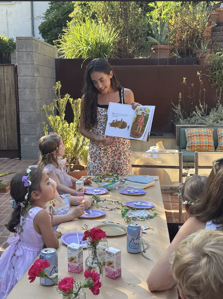 Elena reading a picture book aloud to a group of kids seated around an outdoor table set with craft supplies
