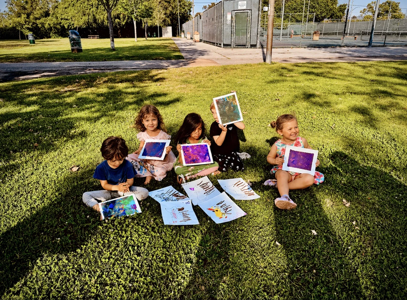 Five toddlers sitting together on the grass at an LA park, holding up their finished marbled paintings with rabbit drawings spread out in front of them