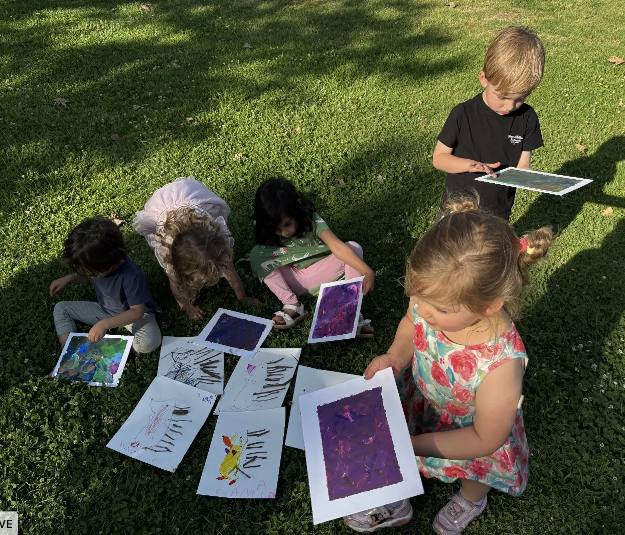 Toddlers in a park sorting through their finished marbled paintings and rabbit drawings laid out on the grass at golden hour