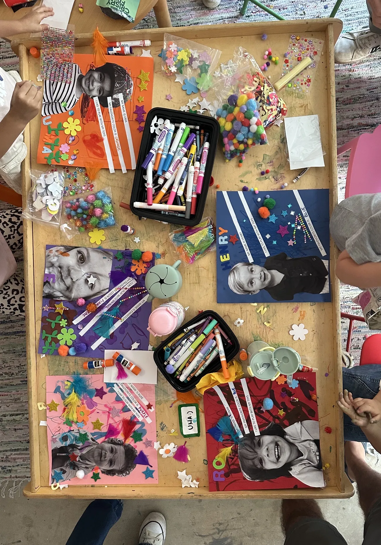Top-down view of a craft table with five 'all about me' self-portrait collages in progress — printed kids' photos decorated with feathers, pom poms, glitter stars, and flower stickers