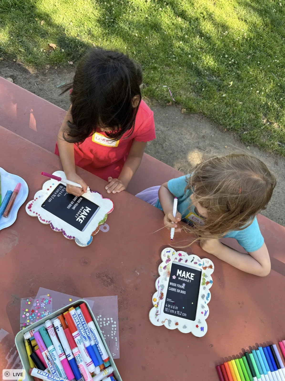 Two girls at a park picnic table working on art-frame projects with markers