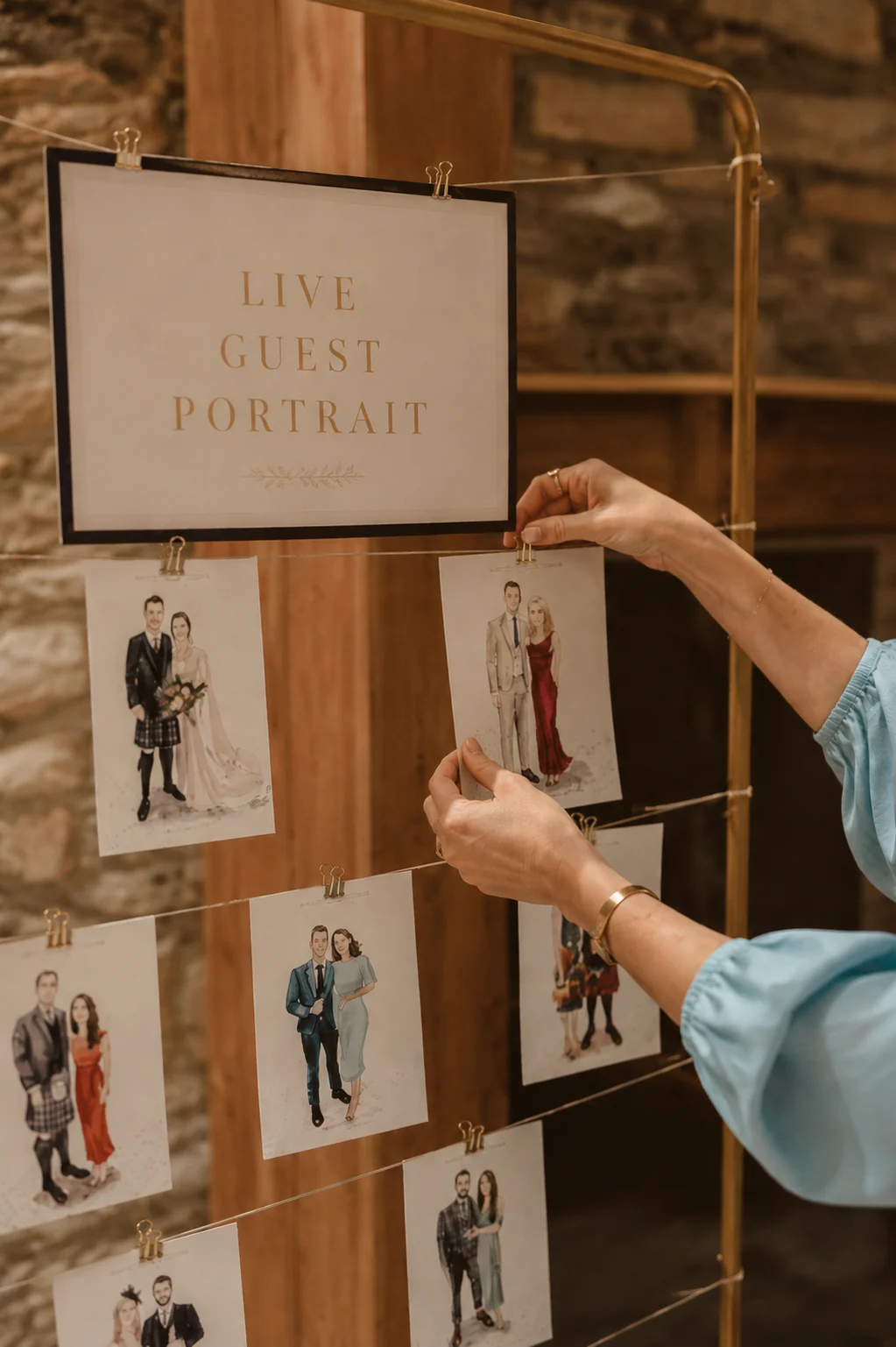 Finished watercolor guest portraits pinned with brass clips to a standing display rack under a Live Guest Portrait sign at a wedding