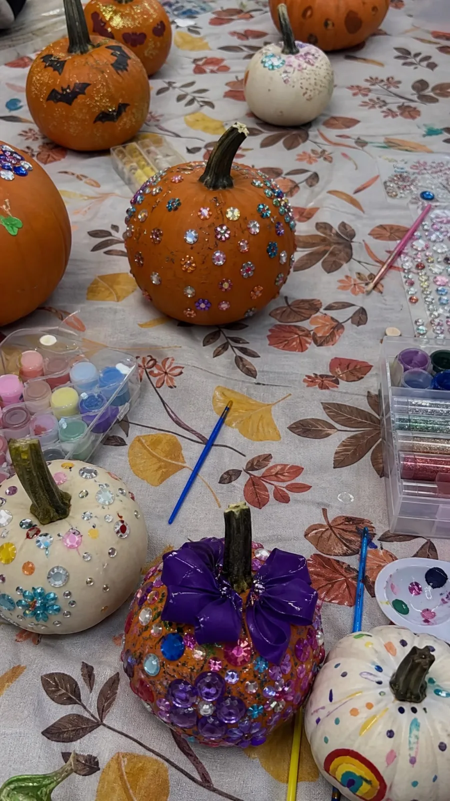 Decorated pumpkins covered in rhinestones, painted bats, and glitter on a fall tablecloth, surrounded by trays of paint, gems, and brushes